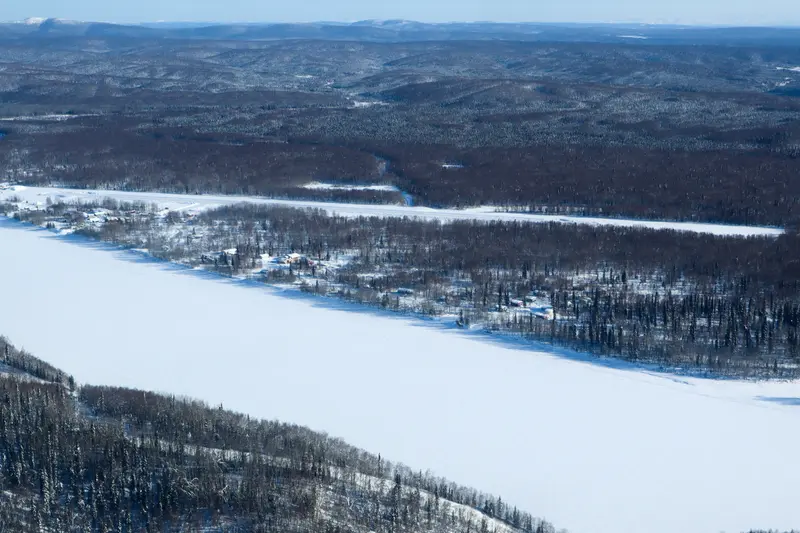 A high view of a wooded area where a few houses are scattered among the trees. On either side of the village are wide, treeless, snow-covered areas where the river is frozen.