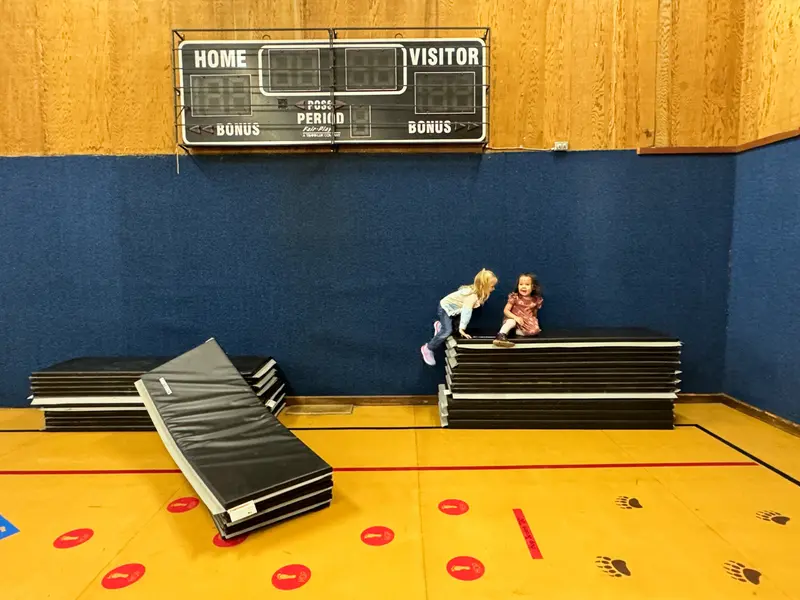 Two children climb on a pile of mats in a gym.