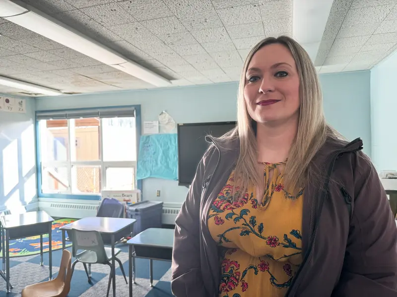 A woman smiles at the camera. Behind her is a sunlit classroom where some of the ceiling panels are stained and sagging.