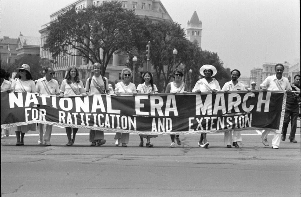 10 women march with a banner that reads "National ERA March for Ratification and Extension." Visible at center is the curved facade of the Federal Trade Commission building and, behind it, the tower of the Old Post Office Pavilion. 