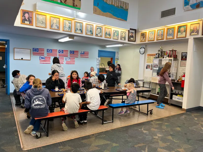 Children eat lunch in a small carpeted area. Framed portraits hang around the upper edge of the room.